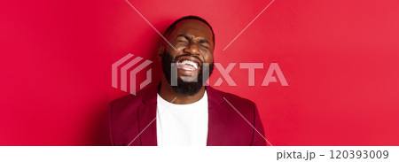 Close-up of happy and carefree Black man having fun, laughing and smiling, standing in blazer against red background 120393009