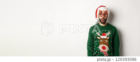 Christmas holidays, celebration concept. Surprised man in Santa hat and party glasses looking at something awesome, standing over white background 120393086