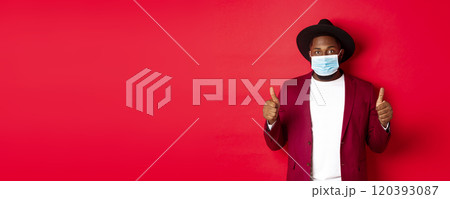 Covid-19 and fashion concept. Stylish african american man in hat and blazer, wearing face mask and showing thumb up, standing over red background 120393087