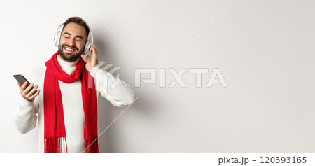 Winter holidays and technology concept. Satisfied man listening music in headphones with closed eyes, smiling with pleasure, holding smartphone, white background Winter holidays and technology concept. Satisfied man listening music in headphones with closed eyes, smiling with pleasure, holding smartphone, white background 120393165