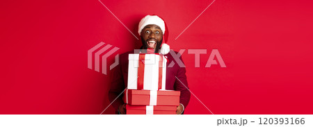 Christmas, New Year and shopping concept. Happy Black man in santa hat and blazer holding xmas presents, bring gifts and smiling, standing against red background Christmas, New Year and shopping concept. Happy Black man in santa hat and blazer holding xmas presents, bring gifts and smiling, standing against red background 120393166