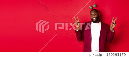 Merry Christmas. Cheerful african american bearded guy in party headband, smiling and showing peace signs, standing over red background 120393170