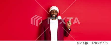 Christmas, party and holidays concept. Handsome Black man in santa hat pointing finger left, showing advertisement, standing happy over red background 120393429