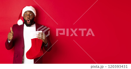 Impressed african american man holding christmas sock with presents, showing thumb up, looking surprised, standing over red background Impressed african american man holding christmas sock with presents, showing thumb up, looking surprised, standing over red background 120393431