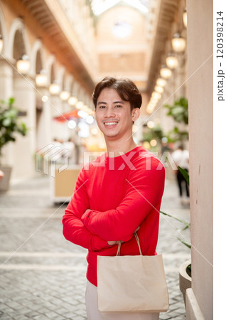 A handsome Asian man carrying a shopping bag, standing in a stunning shopping mall during winter. 120398214