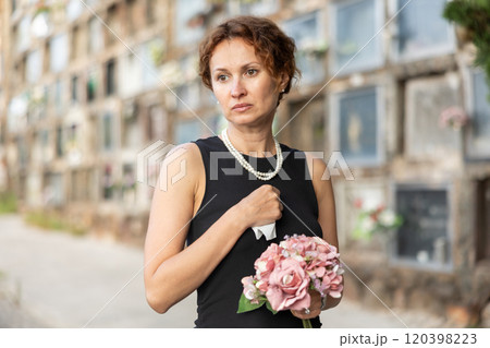 Sad woman in black dress with bouquet of flowers and handkerchief in hands, stands in cemetery Sad woman in black dress with bouquet of flowers and handkerchief in hands, stands in cemetery 120398223