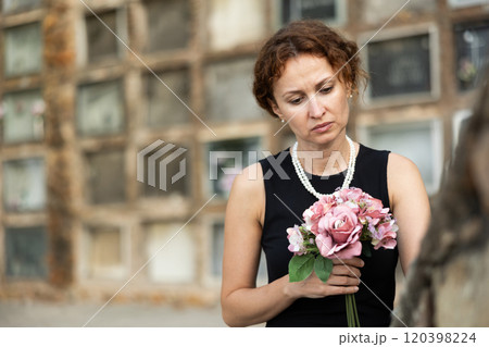 Woman in black dress with bouquet of flowers came to cemetery to honor the memory of deceased loved one 120398224