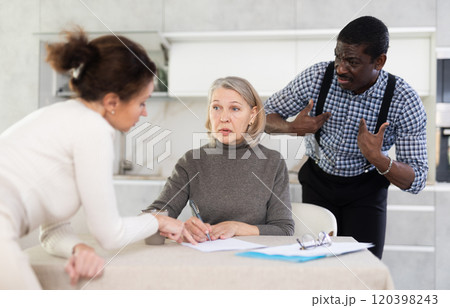 Old woman compiling testament while middle-aged man and woman competing with each other in the kitchen 120398243