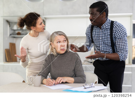 Old woman compiling testament while middle-aged man and woman competing with each other in the kitchen 120398254