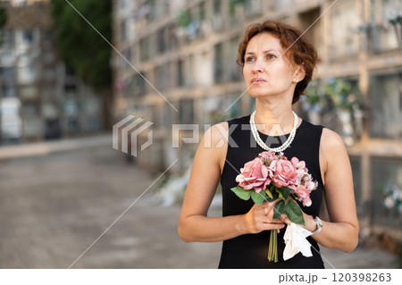 Sad woman in black dress with bouquet of flowers and handkerchief in hands, stands in cemetery 120398263