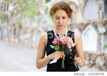Sad woman in black dress with bouquet of flowers and handkerchief in hands, stands in cemetery 120398350