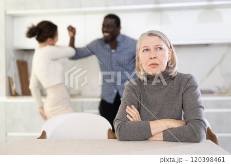 Old woman sitting at the kitchen table with her back to couple quarreling together Old woman sitting at the kitchen table with her back to couple quarreling together 120398461