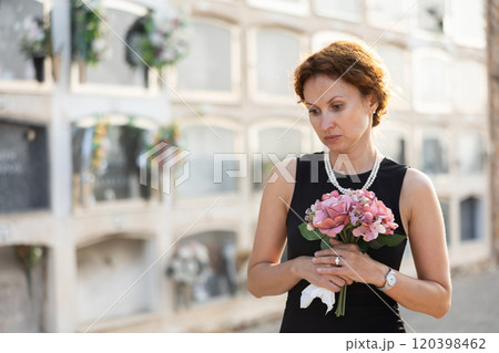 Sad woman in black dress with bouquet of flowers and handkerchief in hands, stands in cemetery Sad woman in black dress with bouquet of flowers and handkerchief in hands, stands in cemetery 120398462