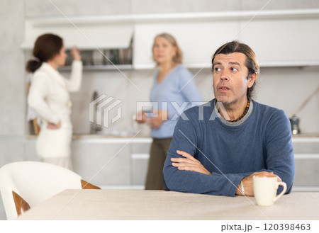 Frustrated man sitting in kitchen during quarrel between his wife and elderly mother. Frustrated man sitting in kitchen during quarrel between his wife and elderly mother. 120398463