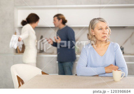 Frustrated senior woman is sitting in kitchen during quarrel between her son and his wife 120398684