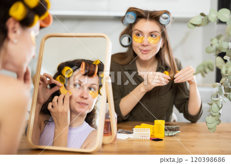 Woman in colorful curlers applying under eye patches by mirror with bestie 120398686