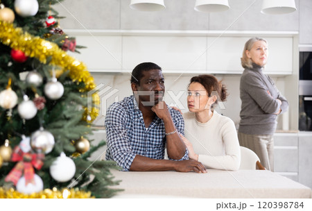 Middle-aged couple sitting at table and mature woman scolding them in the kitchen with Christmas-tree Middle-aged couple sitting at table and mature woman scolding them in the kitchen with Christmas-tree 120398784