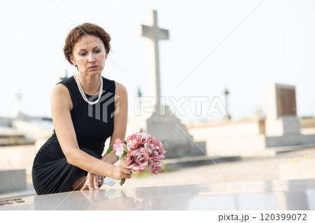 Sorrowful woman puts bouquet of hydrangeas on grave of deceased friend, mourns loss 120399072