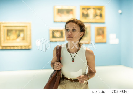 Interested woman looking closely at paintings in art gallery and holding tour sheet in hands Interested woman looking closely at paintings in art gallery and holding tour sheet in hands 120399362