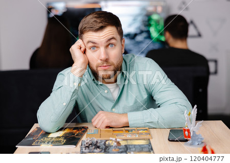 Man with board game piece while practicing strategy in home alone. A man plays a board game with cards and figures, expressing different emotions Man with board game piece while practicing strategy in home alone. A man plays a board game with cards and figures, expressing different emotions 120404777