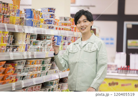 スーパーで買い物中カップラーメンを見せる作業服姿の若い男性 120405519