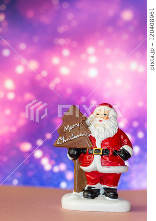 Santa Claus figurine holding a Merry Christmas tree sign, set against a glowing bokeh background in warm festive tones Santa Claus figurine holding a Merry Christmas tree sign, set against a glowing bokeh background in warm festive tones 120408961