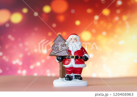 Santa Claus figurine holding a Merry Christmas tree sign, set against a glowing bokeh background in warm festive tones 120408963