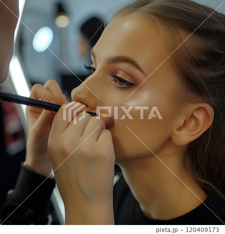 Young beautiful female model doing makeup in a professional studio and looking at the camera with a joyful expression. Makeup artist and hairdresser hold brushes around woman's face. Professional work 120409173
