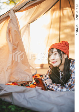 キャンプでテントに寝ながらスマホをいじる女性 120409458