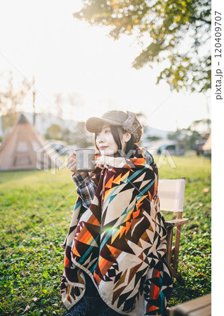 キャンプ場で毛布にくるまりながら飲み物を飲む女性 120409707