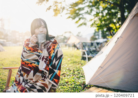 キャンプ場で毛布にくるまりながら飲み物を飲む女性 120409721