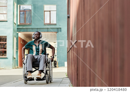 Full length portrait of young African American man with disability using wheelchair and wearing headphones in city moving towards camera, copy space 120418480
