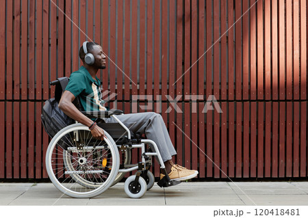 Minimal side view full shot of African American man with disability using wheelchair and wearing headphones listening to music in urban city setting against ribbed metal wall, copy space 120418481