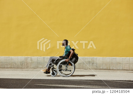 Minimal side view of African American man using wheelchair outdoors in sunlight against vibrant yellow wall copy space 120418504