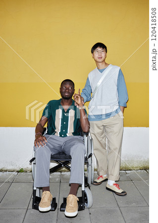 Vertical full length portrait of young African American man with disability posing with friend outdoors against yellow wall shot with flash Vertical full length portrait of young African American man with disability posing with friend outdoors against yellow wall shot with flash 120418508