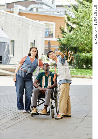 Vertical full length shot of diverse group of young people taking selfie photo outdoors focus on African American man using wheelchair Vertical full length shot of diverse group of young people taking selfie photo outdoors focus on African American man using wheelchair 120418519