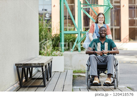 Front view portrait of smiling African American man with disability using wheelchair while moving up on ramp with friend or partner assisting copy space 120418576