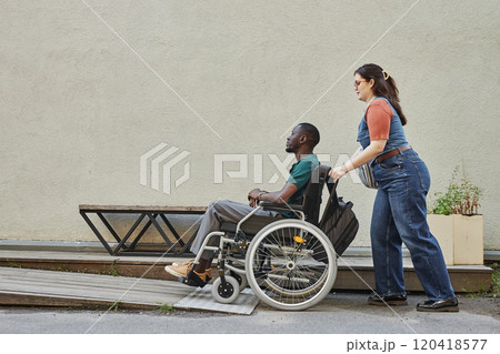 Minimal full length shot of young African American man with disability moving up on ramp with friend or partner assisting copy space Minimal full length shot of young African American man with disability moving up on ramp with friend or partner assisting copy space 120418577