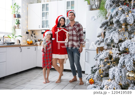Happy family in santa hats with tiny house in cozy home with Christmas decor of festive white kitchen. Gift for New Year. Insurance, moving to new house, mortgage, rent and purchase real estate Happy family in santa hats with tiny house in cozy home with Christmas decor of festive white kitchen. Gift for New Year. Insurance, moving to new house, mortgage, rent and purchase real estate 120419589