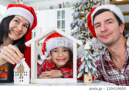 Happy family in santa hats with tiny house in cozy home with Christmas decor of festive white kitchen. Gift for New Year. Insurance, moving to new house, mortgage, rent and purchase real estate 120419600