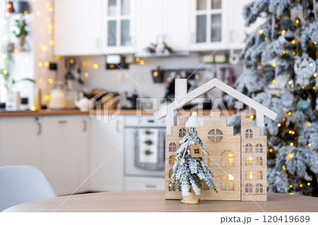 Key on Christmas tree and tiny house on cozy home with Christmas decor on table of festive white kitchen. Gift for New Year. Insurance, moving to new house, mortgage, rent and purchase real estate 120419689
