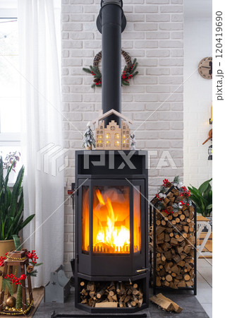 Metall black stove, burning hearth fireplace in white Festive interior of house is decorated for Christmas and New Year, Christmas tree. firewood in the woodpile, cozy and heating of home 120419690