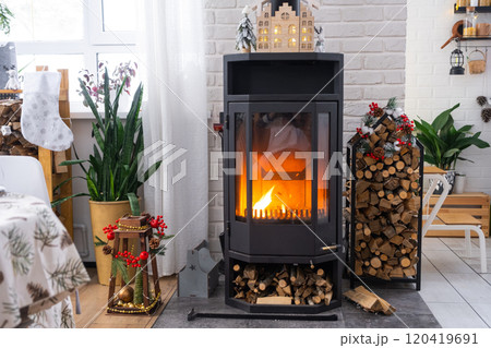 Metall black stove, burning hearth fireplace in white Festive interior of house is decorated for Christmas and New Year, Christmas tree. firewood in the woodpile, cozy and heating of home 120419691