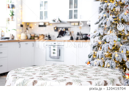 Round empty table to demonstrate the product with with tablecloth with pine needles and cones print, space for text in white kitchen, modern interior with a Christmas tree. New Year, Christmas Round empty table to demonstrate the product with with tablecloth with pine needles and cones print, space for text in white kitchen, modern interior with a Christmas tree. New Year, Christmas 120419704