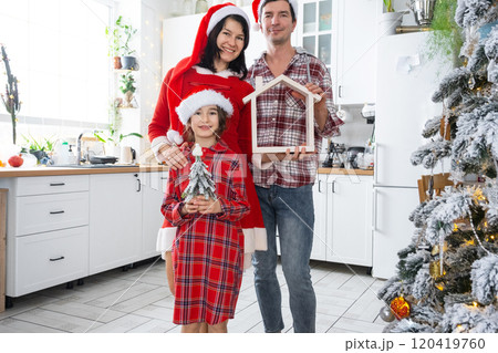 Happy family in santa hats with tiny house in cozy home with Christmas decor of festive white kitchen. Gift for New Year. Insurance, moving to new house, mortgage, rent and purchase real estate 120419760