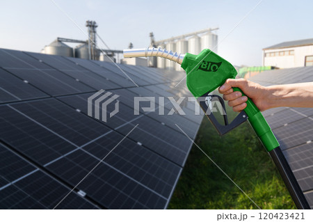 Hand with biofuel refueling nozzle on a background of solar panels and agricultural silos Hand with biofuel refueling nozzle on a background of solar panels and agricultural silos 120423421
