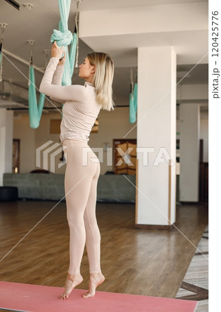 Young woman making antigravity yoga exercises in green hammock at white studio. Professional yoga teacher practicing aerial yoga. Blonde woman wearing beige leggins and shirt. Young woman making antigravity yoga exercises in green hammock at white studio. Professional yoga teacher practicing aerial yoga. Blonde woman wearing beige leggins and shirt. 120425776
