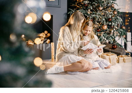 Blonde mother and little daughter sitting near christmas tree at home. Girl with her mother reading a book. Woman and girl wearing white and beige clothes. Blonde mother and little daughter sitting near christmas tree at home. Girl with her mother reading a book. Woman and girl wearing white and beige clothes. 120425808