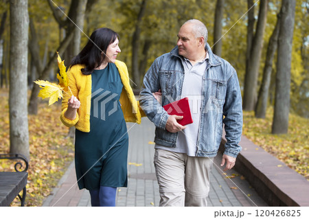 A beautiful middle-aged couple is walking in the autumn park. An elderly man and woman are walking along the road. 120426825