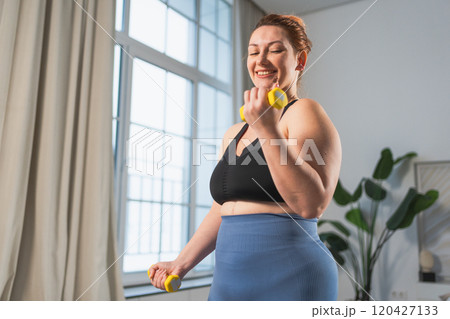 Fitness workout training. Woman picking up dumbbells. Plus size female doing sports exercise at home indoor. Girl in sportswear have workout weight training for weight loss. Sport and fitness Fitness workout training. Woman picking up dumbbells. Plus size female doing sports exercise at home indoor. Girl in sportswear have workout weight training for weight loss. Sport and fitness 120427133